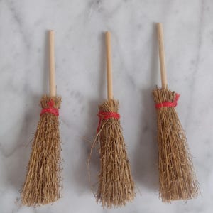 May include: Three miniature brooms with natural straw bristles and wooden handles. Each broom is tied with red twine. The brooms are arranged in a row against a white marble background.