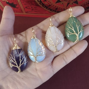 May include: Four teardrop-shaped gemstone pendants with gold wire tree designs. The gemstones are purple, light blue, pink, and green. Each pendant has a gold loop for hanging. The pendants are displayed on a hand.