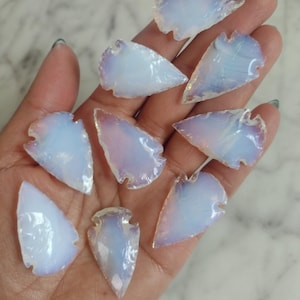 May include: A collection of opalescent, arrowhead-shaped stones. The stones have a translucent, milky white appearance with subtle iridescent hues of blue and pink. The edges are roughly chipped, giving them a natural, handcrafted look.