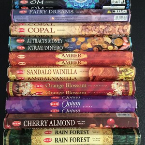 May include: A stack of 12 incense sticks boxes with various scents, including Lotus, Om, Fairy Dreams, Copal, Attracts Money, Amber, Sandal Vanilla, Orange Blossom, Opium, Cherry Almond, Rain Forest, and Jasmine Blossom. The boxes are all in a similar style, with a white background and black text. The boxes are stacked on a black surface.