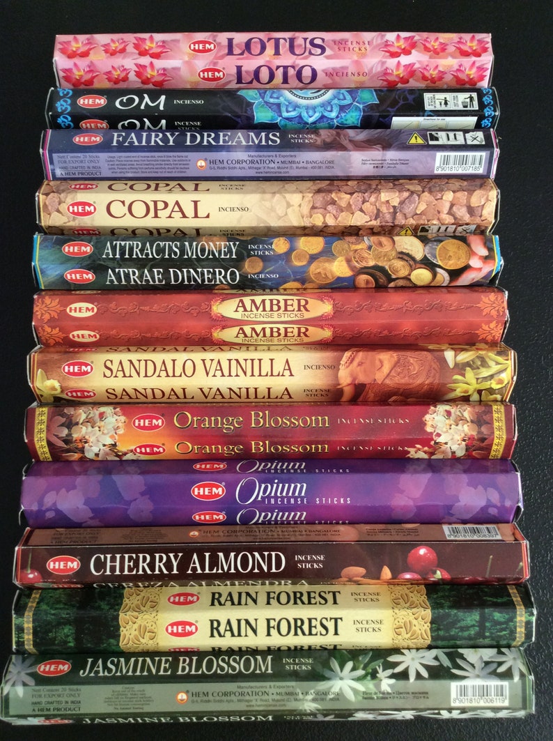 May include: A stack of 12 incense sticks boxes with various scents, including Lotus, Om, Fairy Dreams, Copal, Attracts Money, Amber, Sandal Vanilla, Orange Blossom, Opium, Cherry Almond, Rain Forest, and Jasmine Blossom. The boxes are all in a similar style, with a white background and black text. The boxes are stacked on a black surface.