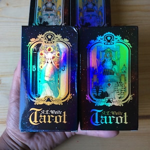 May include: Two black boxes with holographic foil designs featuring the text "A.E. Waite Tarot" in gold lettering. The boxes contain tarot cards.
