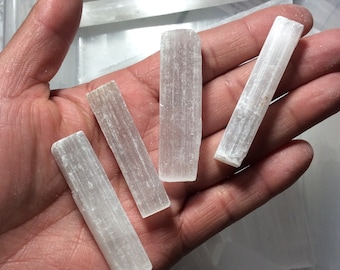 May include: Four white selenite sticks of varying lengths, held in a hand. The sticks are smooth and have a striated texture.