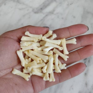 May include: A collection of small, off-white bone-shaped objects. The bones are clustered together, with a few scattered across the palm. The bones have a detailed, realistic appearance, suggesting they are miniature replicas or decorative items.