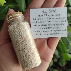 May include: A small glass bottle filled with white star sand from Okinawa, Japan. The bottle has a cork stopper and is held in a hand. The label reads "Star Sand From Okinawa, Japan Star sand is a biogenic sand made of skeletons of tiny, mono-celled sea creatures belonging to the foraminifera group. Used to promote happiness and carried as a lucky talisman."
