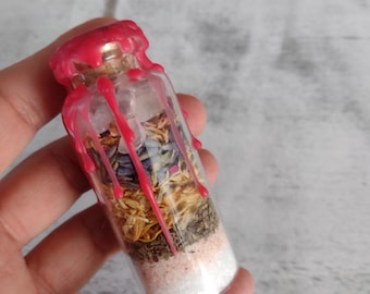 May include: A small glass bottle filled with dried herbs, flowers, and salt, sealed with a red wax top. The bottle is held in a hand.