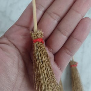 May include: Miniature brooms with natural straw bristles and wooden handles. The brooms are tied with red twine. The brooms are small, likely for decorative purposes or crafts.