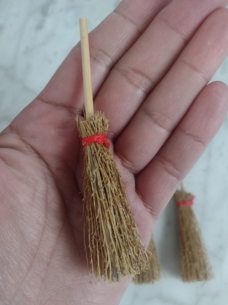 May include: Miniature brooms with natural straw bristles and wooden handles. The brooms are tied with red twine. The brooms are small, likely for decorative purposes or crafts.