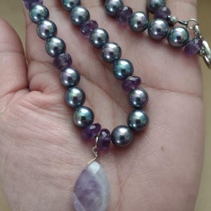 May include: A necklace featuring dark gray pearls and small purple amethyst beads. A teardrop-shaped amethyst pendant hangs from the necklace. The necklace is displayed against a neutral background.