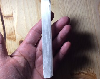 May include: A white selenite wand, also known as a cleansing wand, is held in a person's hand. The wand is smooth and has a natural, striated texture.