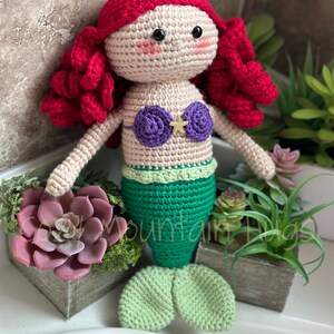 Puede incluir: Una mu&ntilde;eca de sirena de crochet con cabello rojo, un sujetador de concha p&uacute;rpura y una cola verde. La mu&ntilde;eca est&aacute; sentada sobre una superficie blanca con una planta suculenta en maceta en el fondo.