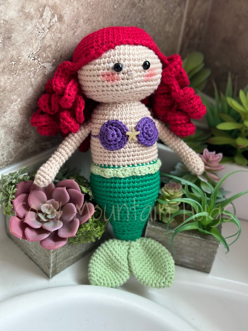 Puede incluir: Una mu&ntilde;eca de sirena de crochet con cabello rojo, un sujetador de concha p&uacute;rpura y una cola verde. La mu&ntilde;eca est&aacute; sentada sobre una superficie blanca con una planta suculenta en maceta en el fondo.