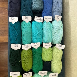 Puede incluir: Una colecci&oacute;n de ovillos de lana en varios tonos de azul, verde y turquesa. Los ovillos est&aacute;n etiquetados con sus nombres de color, incluyendo Azul marino, Denim, Denim claro, Azul real, Azul polvoriento, Azul oscuro, Azul claro, Turquesa medio, Aqua medio, Aqua claro, Verde bosque, Verde hierba, Verde Kelly, Verde primavera, Verde claro, Verde lima, Lima brillante, Menta y Menta claro.