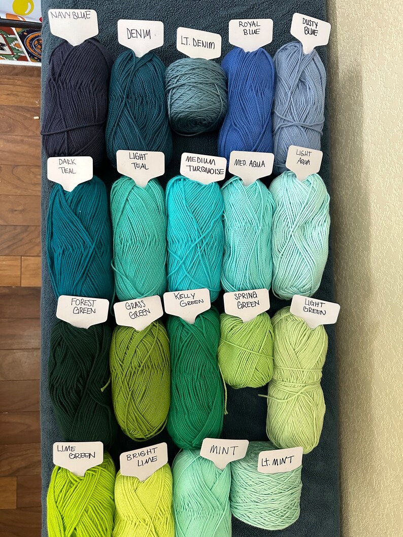 Puede incluir: Una colecci&oacute;n de ovillos de lana en varios tonos de azul, verde y turquesa. Los ovillos est&aacute;n etiquetados con sus nombres de color, incluyendo Azul marino, Denim, Denim claro, Azul real, Azul polvoriento, Azul oscuro, Azul claro, Turquesa medio, Aqua medio, Aqua claro, Verde bosque, Verde hierba, Verde Kelly, Verde primavera, Verde claro, Verde lima, Lima brillante, Menta y Menta claro.