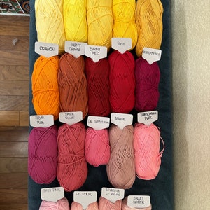 Puede incluir: Una colecci&oacute;n de ovillos de lana en varios tonos de amarillo, naranja, rojo, rosa y morado. Cada ovillo est&aacute; etiquetado con el nombre de su color, como "Amarillo claro", "Amarillo soleado", "Naranja quemado", "Rojo", "Ciruela Subar", "Rosa oscuro", "Rosa beb&eacute;" y "Zapato de ballet".