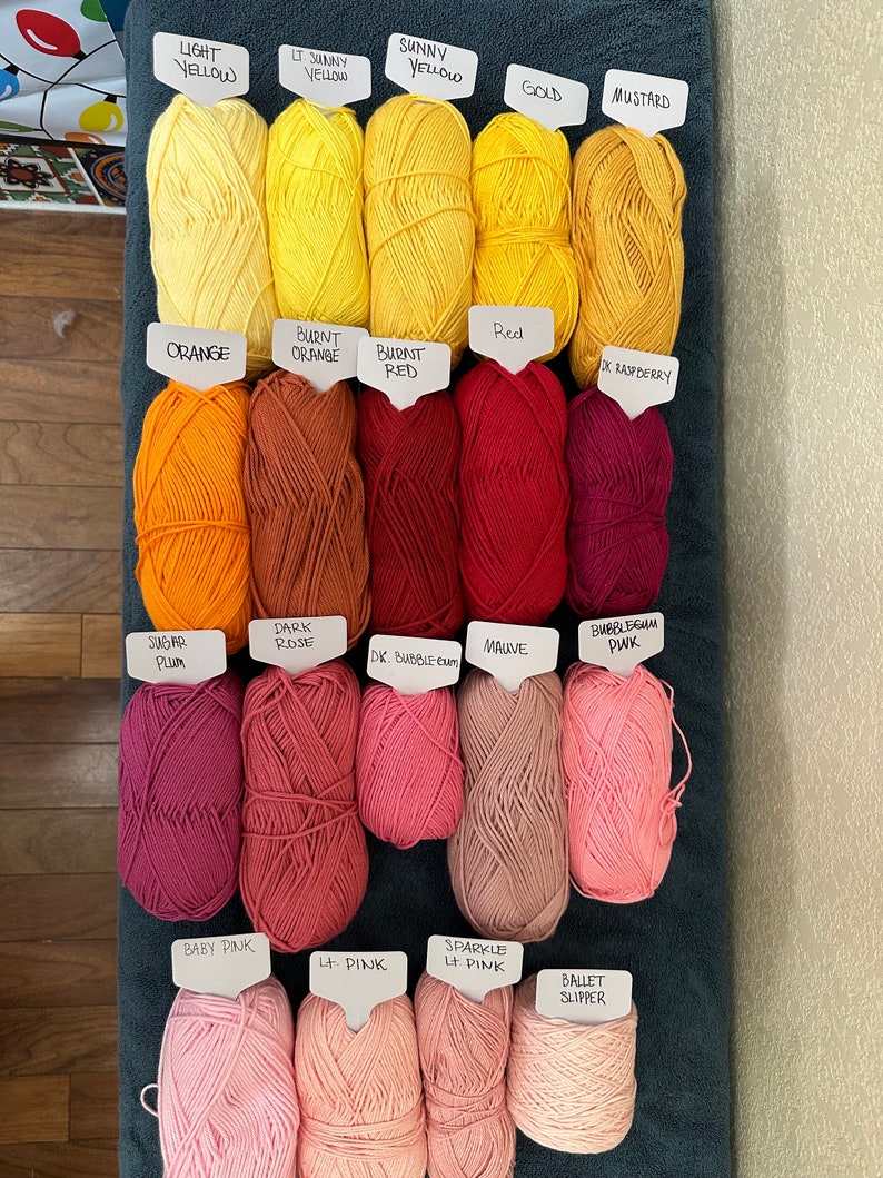Puede incluir: Una colecci&oacute;n de ovillos de lana en varios tonos de amarillo, naranja, rojo, rosa y morado. Cada ovillo est&aacute; etiquetado con el nombre de su color, como "Amarillo claro", "Amarillo soleado", "Naranja quemado", "Rojo", "Ciruela Subar", "Rosa oscuro", "Rosa beb&eacute;" y "Zapato de ballet".