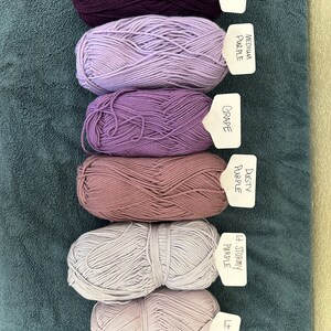 Puede incluir: Seis ovillos de lana en varios tonos de morado, desde morado oscuro a morado claro. Cada ovillo tiene una etiqueta con el nombre del color escrito.