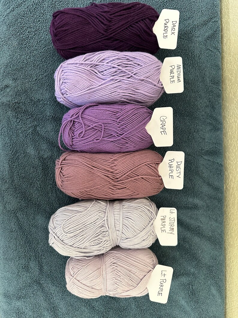 Puede incluir: Seis ovillos de lana en varios tonos de morado, desde morado oscuro a morado claro. Cada ovillo tiene una etiqueta con el nombre del color escrito.