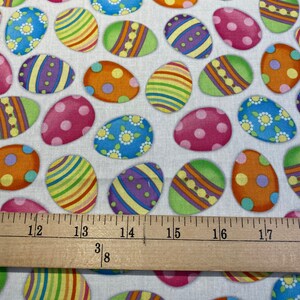May include: White fabric with a repeating pattern of colorful Easter eggs. The eggs are decorated with stripes, polka dots, and floral designs.