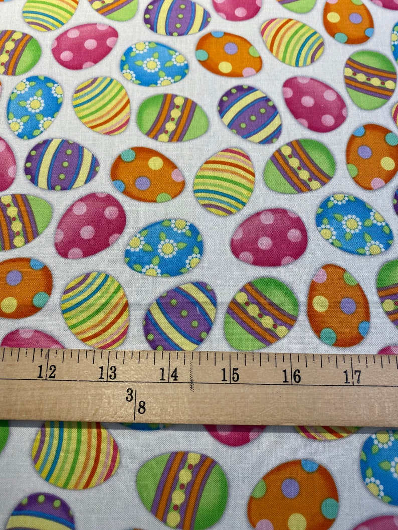 May include: White fabric with a repeating pattern of colorful Easter eggs. The eggs are decorated with stripes, polka dots, and floral designs.