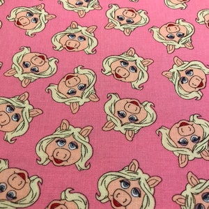 May include: Pink fabric with a repeating pattern of Miss Piggy's face. The fabric features a light pink background with a white outline around each image.