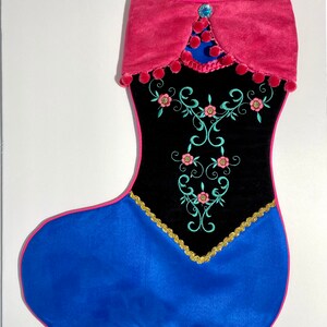 May include: A black velvet Christmas stocking with pink and blue accents. The stocking features embroidered floral designs and a blue jewel at the top.
