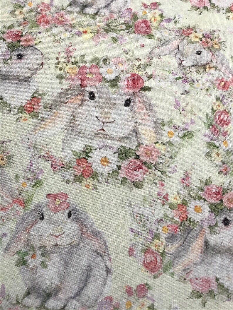 May include: A light green fabric with a repeating pattern of gray bunnies wearing floral crowns. The bunnies are surrounded by pink, white, and purple flowers.