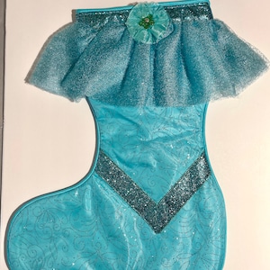 May include: A turquoise blue Christmas stocking with a glittery design and a tulle ruffle at the top. A green flower with a rhinestone center is attached to the ruffle.