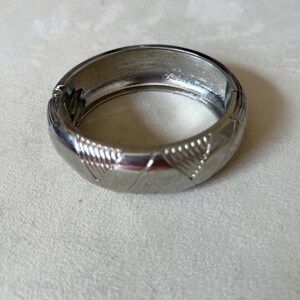 Può includere: Un braccialetto a cerchio incernierato color argento. Il braccialetto ha una forma arrotondata con un motivo strutturato di linee diagonali e forme a diamante. La chiusura è visibile sul lato. Gioielli.