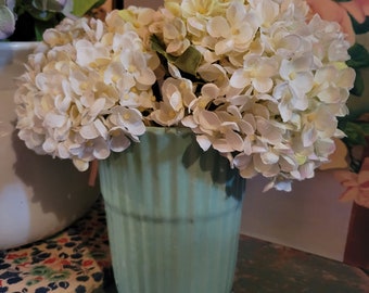 May include: A light green, ribbed vase filled with white and light yellow hydrangea flowers. The vase sits on a distressed, teal-colored wooden surface. The background includes a floral painting and other decorative items.