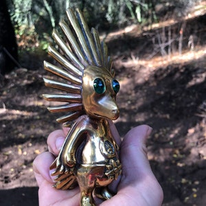 May include: A small, gold-coloured figurine with a spiky, sun-like head and green eyes. The figurine is standing on a hand.