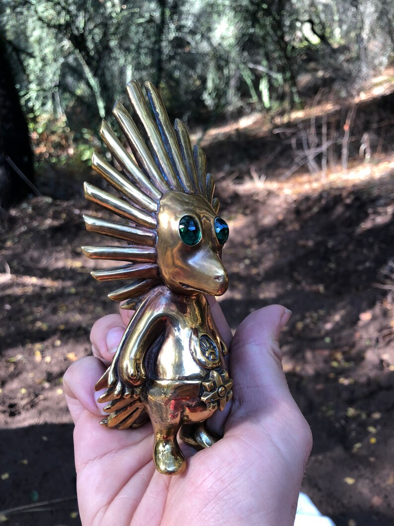 May include: A small, gold-coloured figurine with a spiky, sun-like head and green eyes. The figurine is standing on a hand.