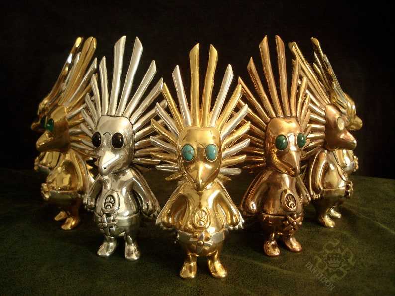 May include: A group of five silver, gold, and bronze figurines with turquoise and black eyes. Each figurine has a spiky crown and a bird-like beak. The figurines are arranged in a circle.
