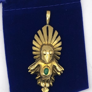 May include: A gold pendant in the shape of a stylised bird with a green gemstone in its chest. The pendant is hanging from a blue velvet pouch.