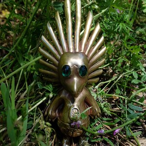 May include: A gold-coloured figurine with a spiky crown and green eyes. The figurine is standing on a bed of brown pine needles and green grass.