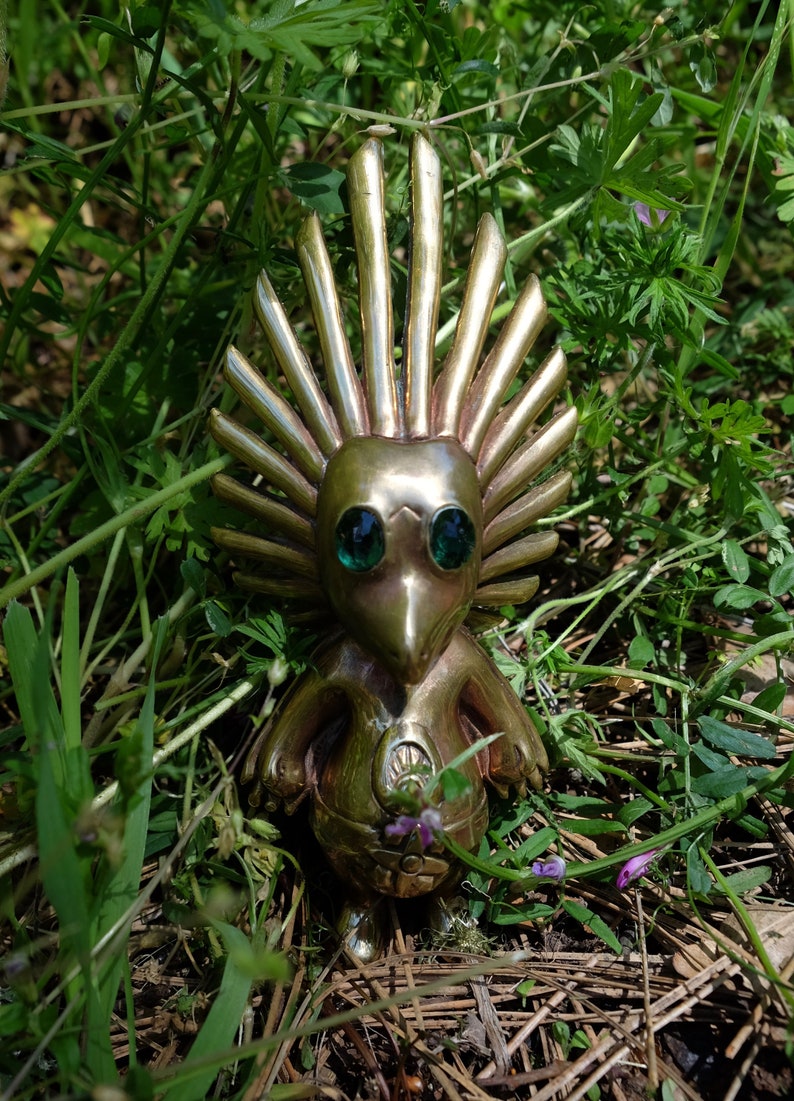 May include: A gold-coloured figurine with a spiky crown and green eyes. The figurine is standing on a bed of brown pine needles and green grass.