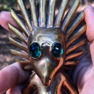 May include: A gold-coloured figurine with a spiky head and green gem eyes. The figurine is wearing a decorative garment with a sun-like symbol on the chest.