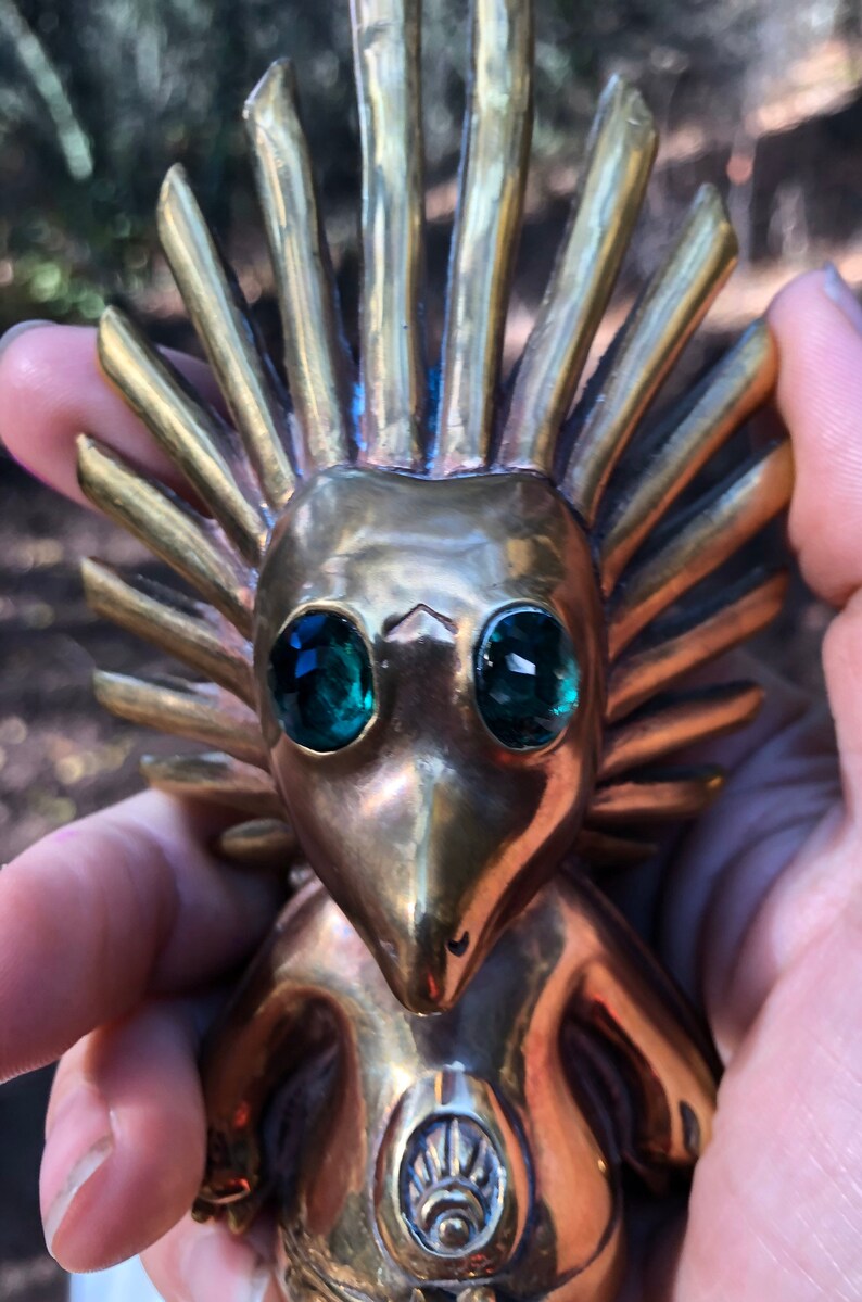 May include: A gold-coloured figurine with a spiky head and green gem eyes. The figurine is wearing a decorative garment with a sun-like symbol on the chest.