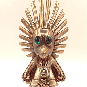 May include: A gold-coloured figurine of a creature with a spiky head, green eyes, and a star-shaped design on its chest. The figurine is standing on a white surface.