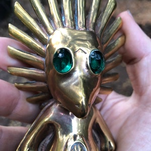 May include: A gold-coloured figurine with a spiky, sun-like head and green gem eyes. The figurine has a stylised human-like body with a circular design on its chest.