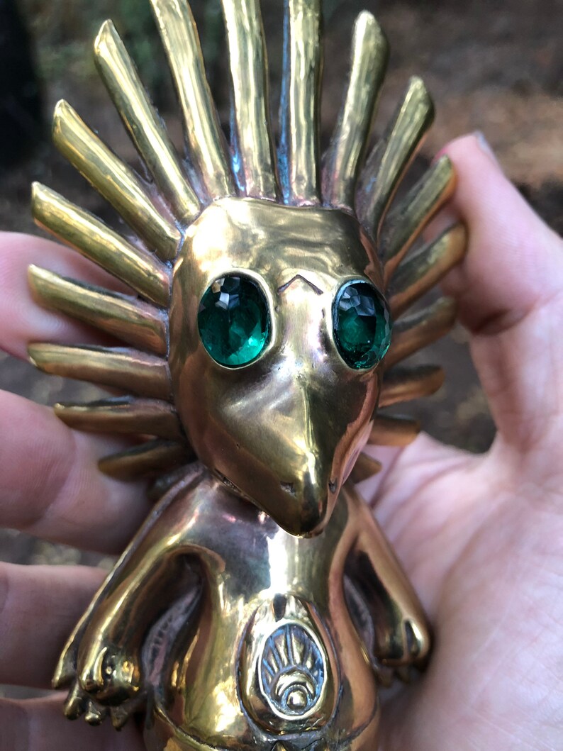 May include: A gold-coloured figurine with a spiky, sun-like head and green gem eyes. The figurine has a stylised human-like body with a circular design on its chest.
