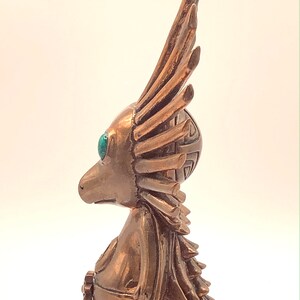 May include: A bronze figurine of a bird-like creature with a long, feathery crest and turquoise eyes. The figurine is standing on a white surface.