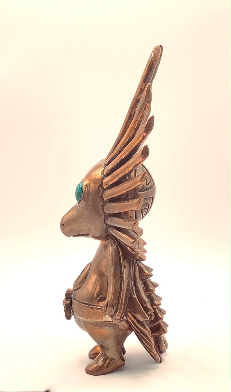 May include: A bronze figurine of a bird-like creature with a long, feathery crest and turquoise eyes. The figurine is standing on a white surface.