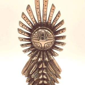 May include: A gold-coloured metal sculpture of a stylised figure with a circular head and a flowing, layered skirt. The head features a geometric design with a cross in the centre.