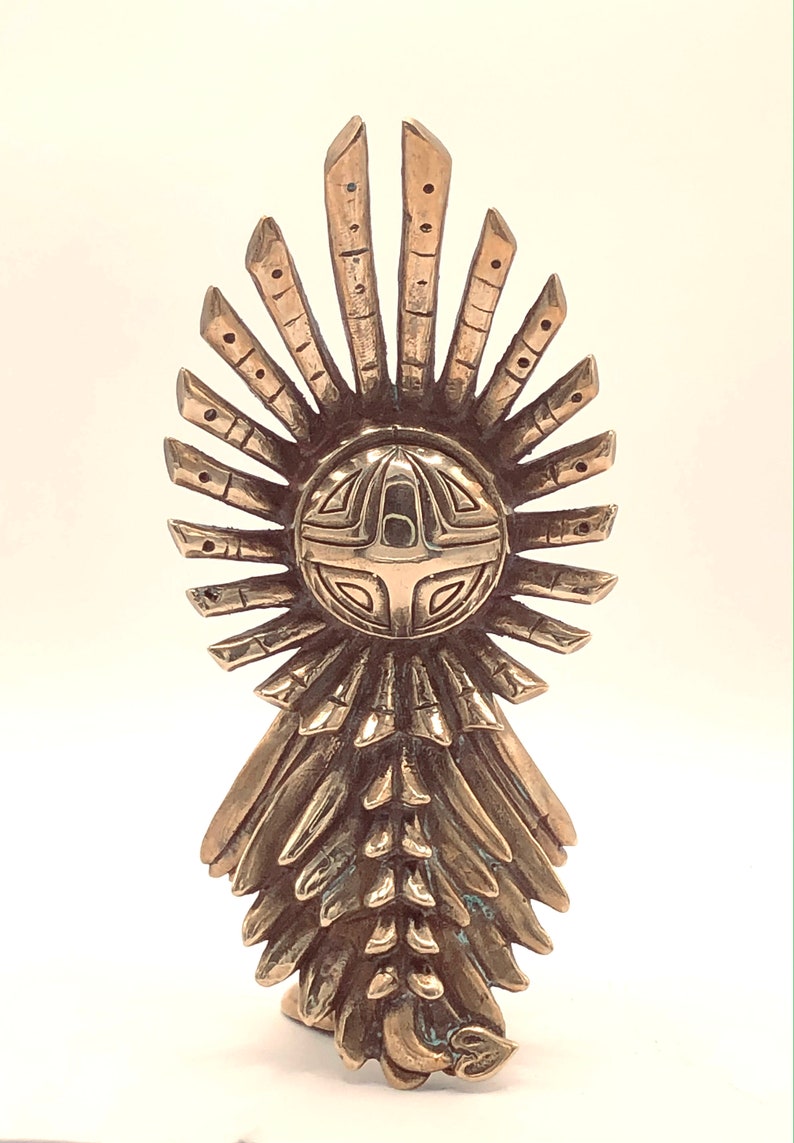 May include: A gold-coloured metal sculpture of a stylised figure with a circular head and a flowing, layered skirt. The head features a geometric design with a cross in the centre.