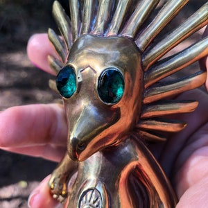 May include: A gold-coloured figurine with a spiky, sun-like head and green eyes. The figurine is holding its hands in front of its chest, and there is a symbol on its chest.