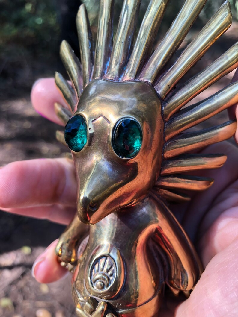 May include: A gold-coloured figurine with a spiky, sun-like head and green eyes. The figurine is holding its hands in front of its chest, and there is a symbol on its chest.