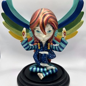 May include: A colourful, hand-painted figurine of a person with wings, sitting in a meditative pose. The figurine is wearing a blue and green striped outfit with white polka dots and has red and gold hair. The wings are multi-coloured and have a rainbow effect.