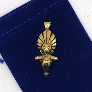 May include: A gold-coloured pendant with an abstract design, featuring a stylised figure with a headdress and outstretched arms. The pendant is suspended against a dark blue velvet pouch.