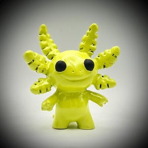 May include: A vibrant yellow axolotl figure with black eyes and a smiling face. The figurine features detailed gills and limbs, and stands upright. The design is cartoon-like and playful, suitable for display as a decorative item.